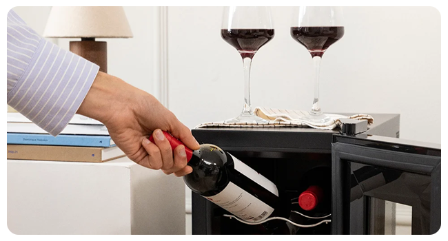 Una mano che aziona un apribottiglie da vino su un supporto nero, con due bicchieri da vino contenenti vino rosso visibili sullo sfondo