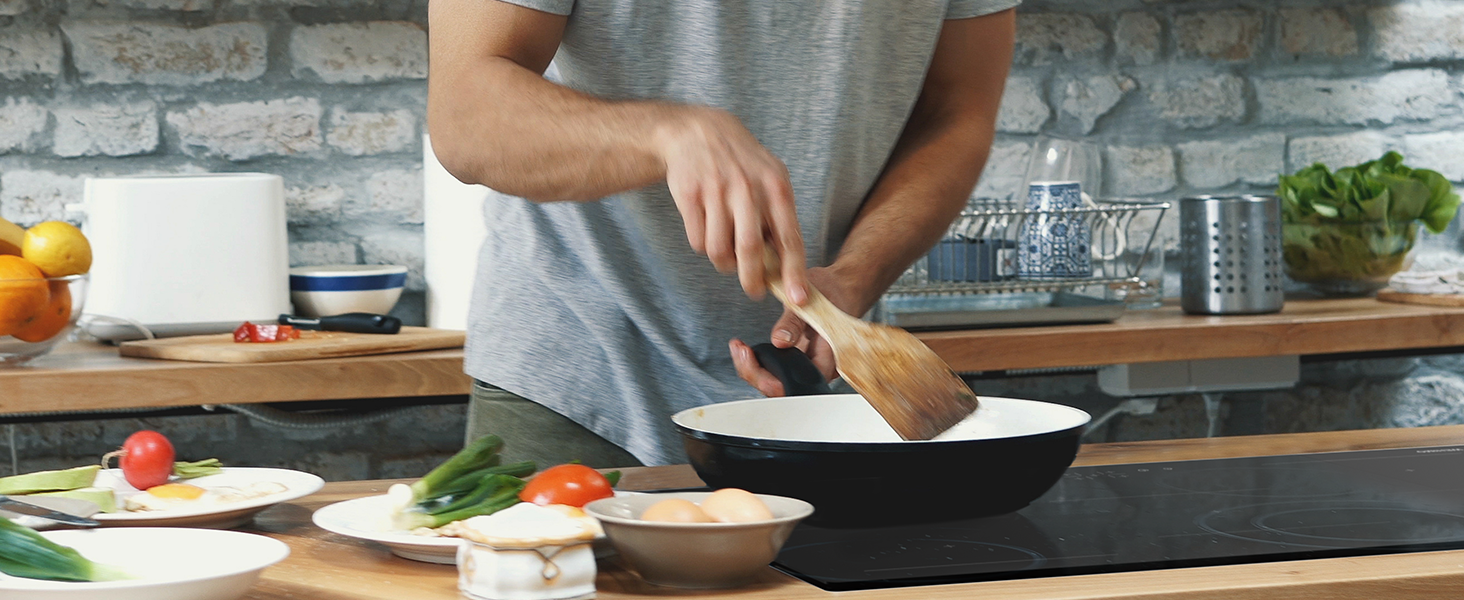 Scena di cucina che mostra le mani usando un utensile di legno con padella bianca su un piano di lavoro scuro, circondate da ingredienti freschi e