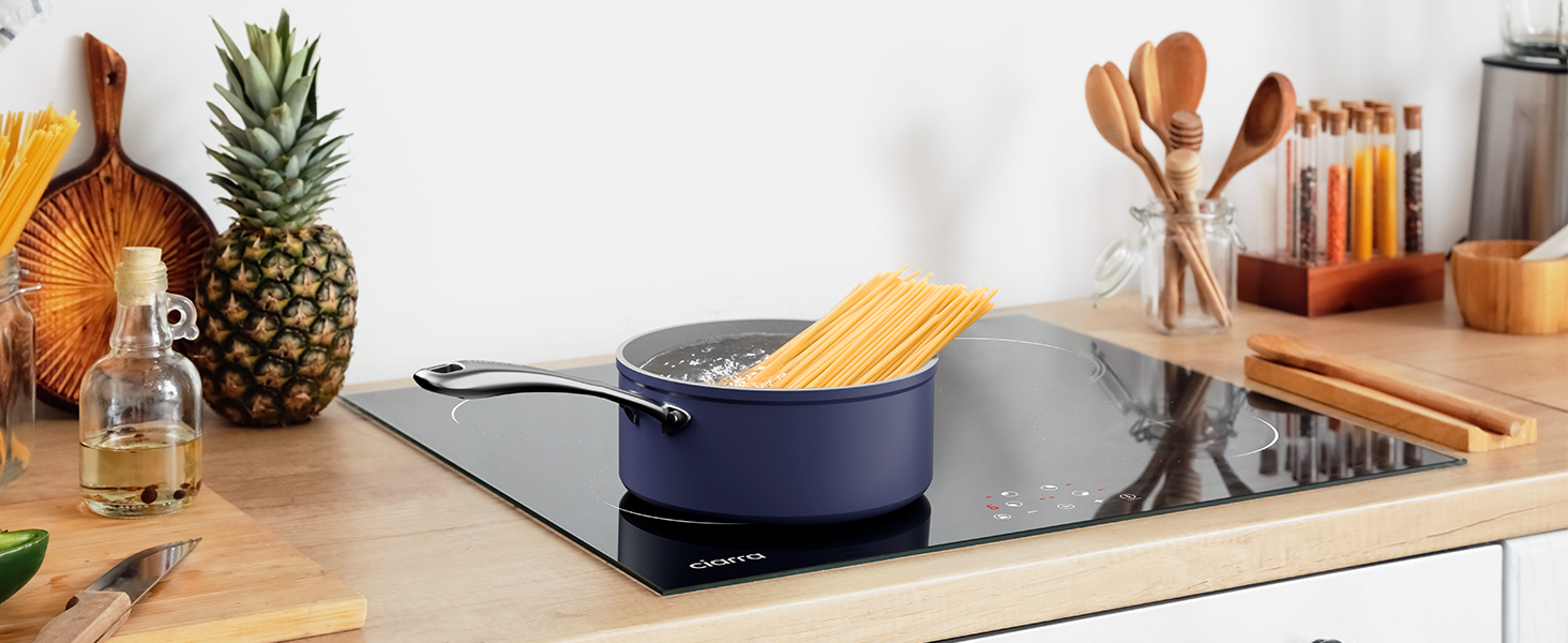 Piano cucina moderno con piano cottura a induzione, pentola blu e vari utensili da cucina. Ananas e altri oggetti visibili sullo sfondo.