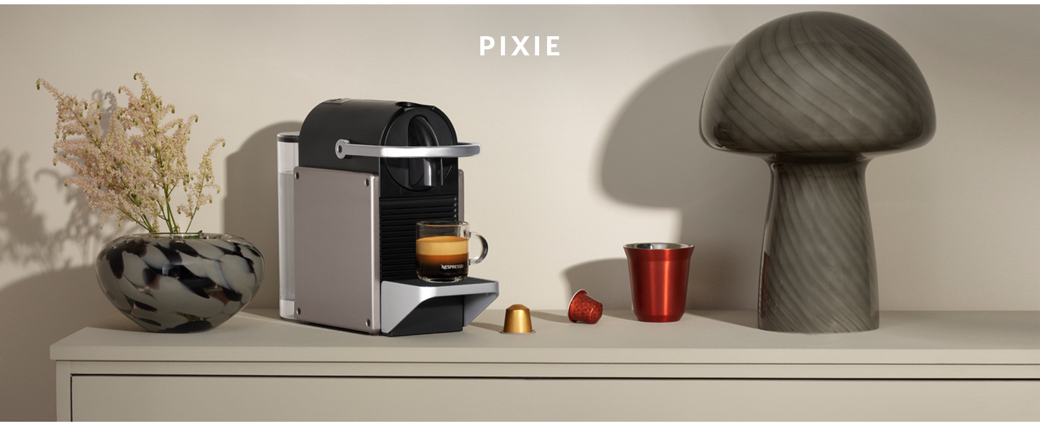 Il testo dice ?PIXIE?. Macchina da caffè con finitura argentata su superficie beige con lampada decorativa a forma di fungo e accessori per il caffè.