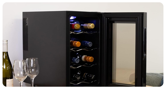 Cantinetta frigo compatta nera con porta in vetro, che espone più bottiglie di vino su scaffali in metallo. Bicchieri da vino e bottiglia visibili in primo piano
