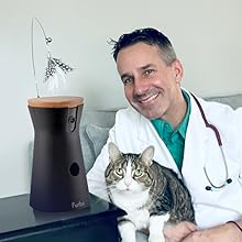 Dispositivo cilindrico nero con giocattolo in piume, simile a un giocattolo interattivo per animali domestici. Gatto soriano e persona in abbigliamento medico visibili nelle vicinanze