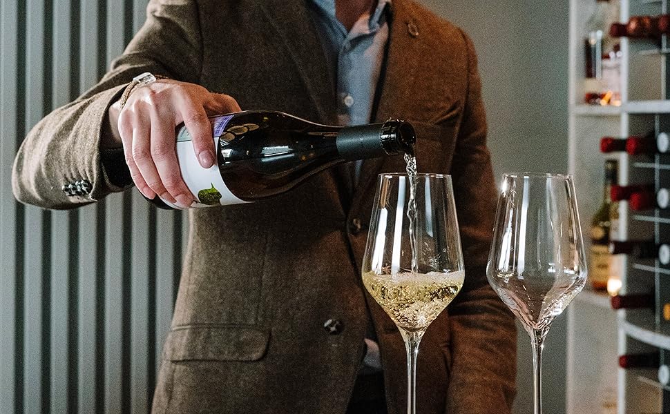 Persona in giacca marrone che versa vino bianco in due bicchieri con stelo, con bottiglie di vino visibili sullo